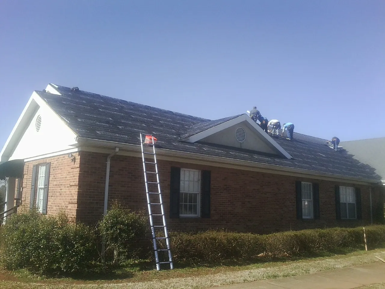 Crew tearing shingles off a brick ranch roof with a ladder set — Emergency Roof Repair work by our crew in Twin Bridges