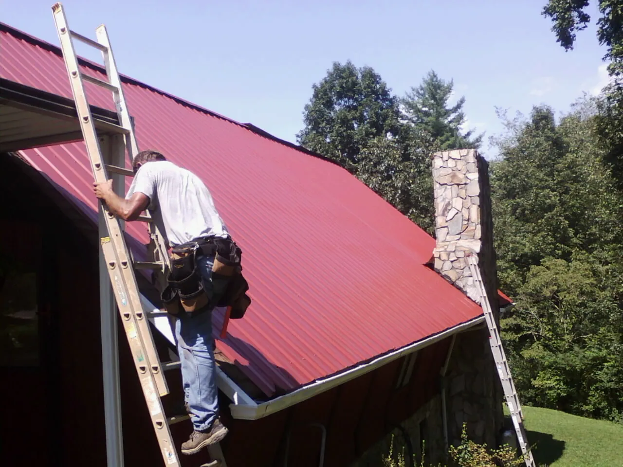 Roofer on a ladder beside a red metal roof and stone chimney — Soffit & Fascia Repair by Roofing Brothers in Twin Bridges